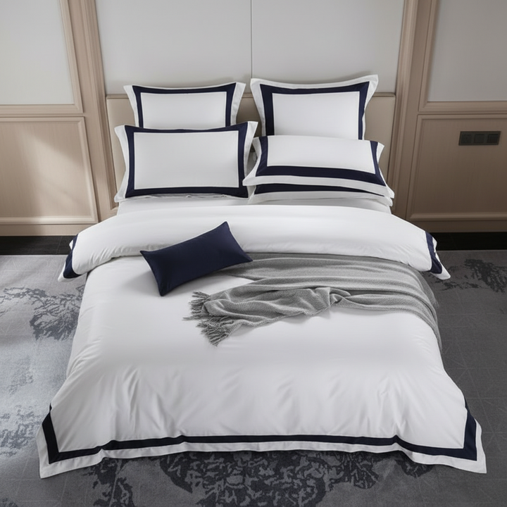 A neatly made bed in a styled bedroom setting, featuring a white duvet cover and multiple pillows with dark navy blue borders and a matching navy accent pillow. The room has beige wood paneling on the walls and a gray patterned rug on the floor.